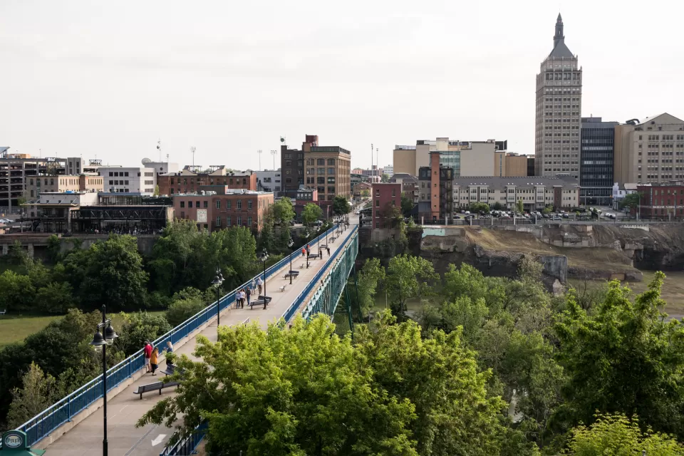 A long pedestrian bridge over a river in the High Falls District with skyline of Rochester, New York, in the backround.